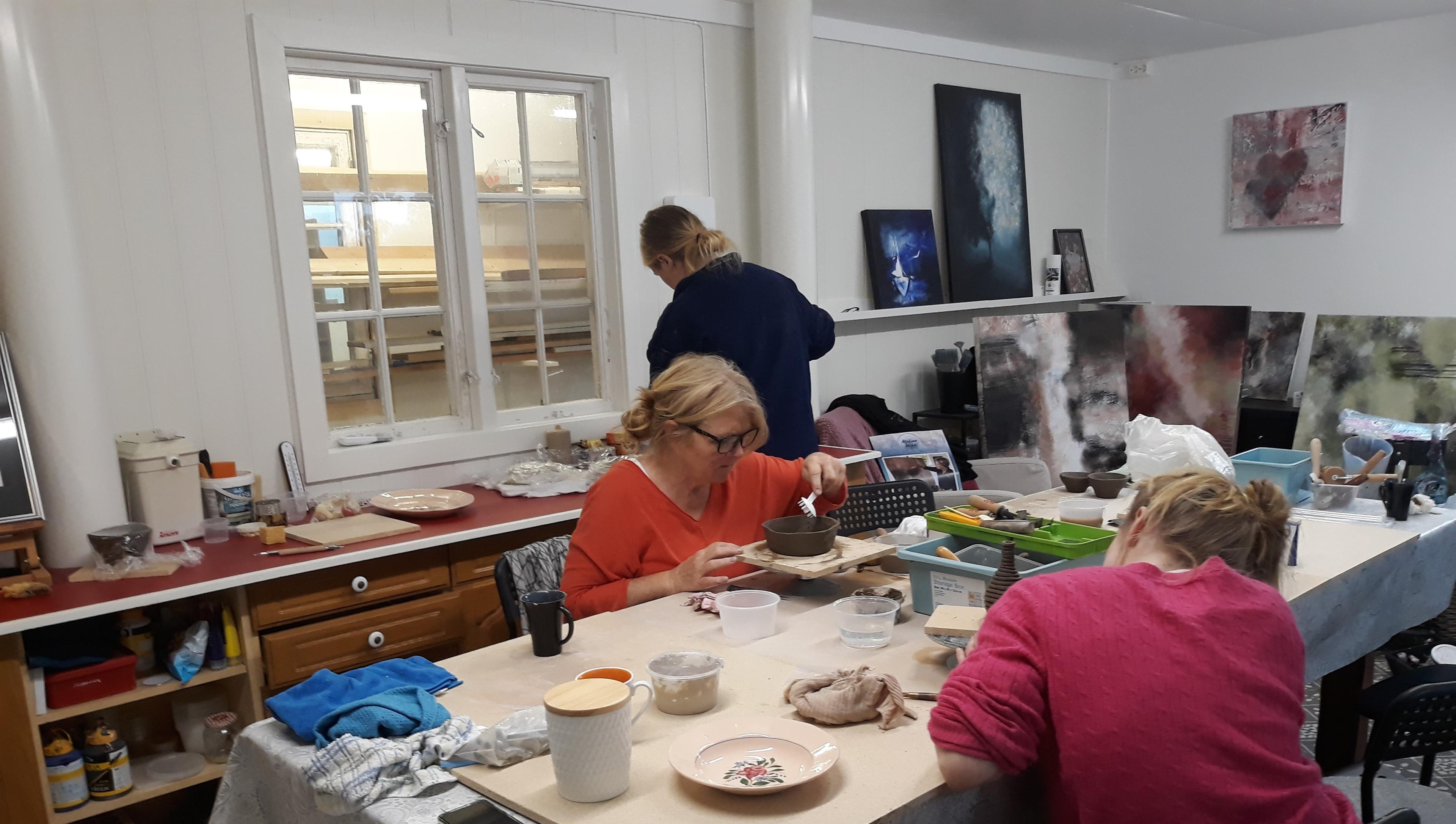 Kunstnere jobber med keramikk og maleri i et lyst atelier med flere arbeidsstasjoner.