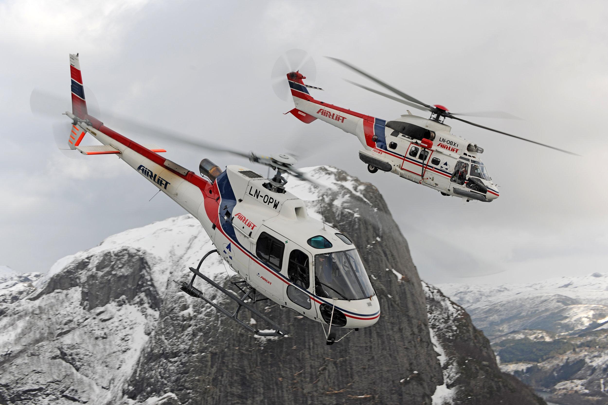 To helikoptre flyr over en snødekt fjelltoppe i et dramatisk landskap.