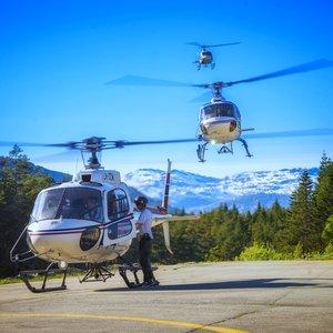 Tre helikoptre på en flyplass med fjell i bakgrunnen og en pilot som forbereder avgang.