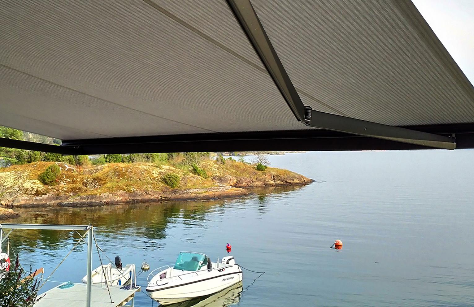 Utsikt fra en terrasse med markise over en båt ved en rolig innsjø og en grønn øy i bakgrunnen.