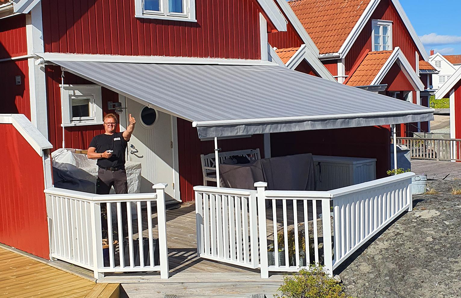 En mann står på en terrasse med et grått markise foran et rødt hus med hvite detaljer.