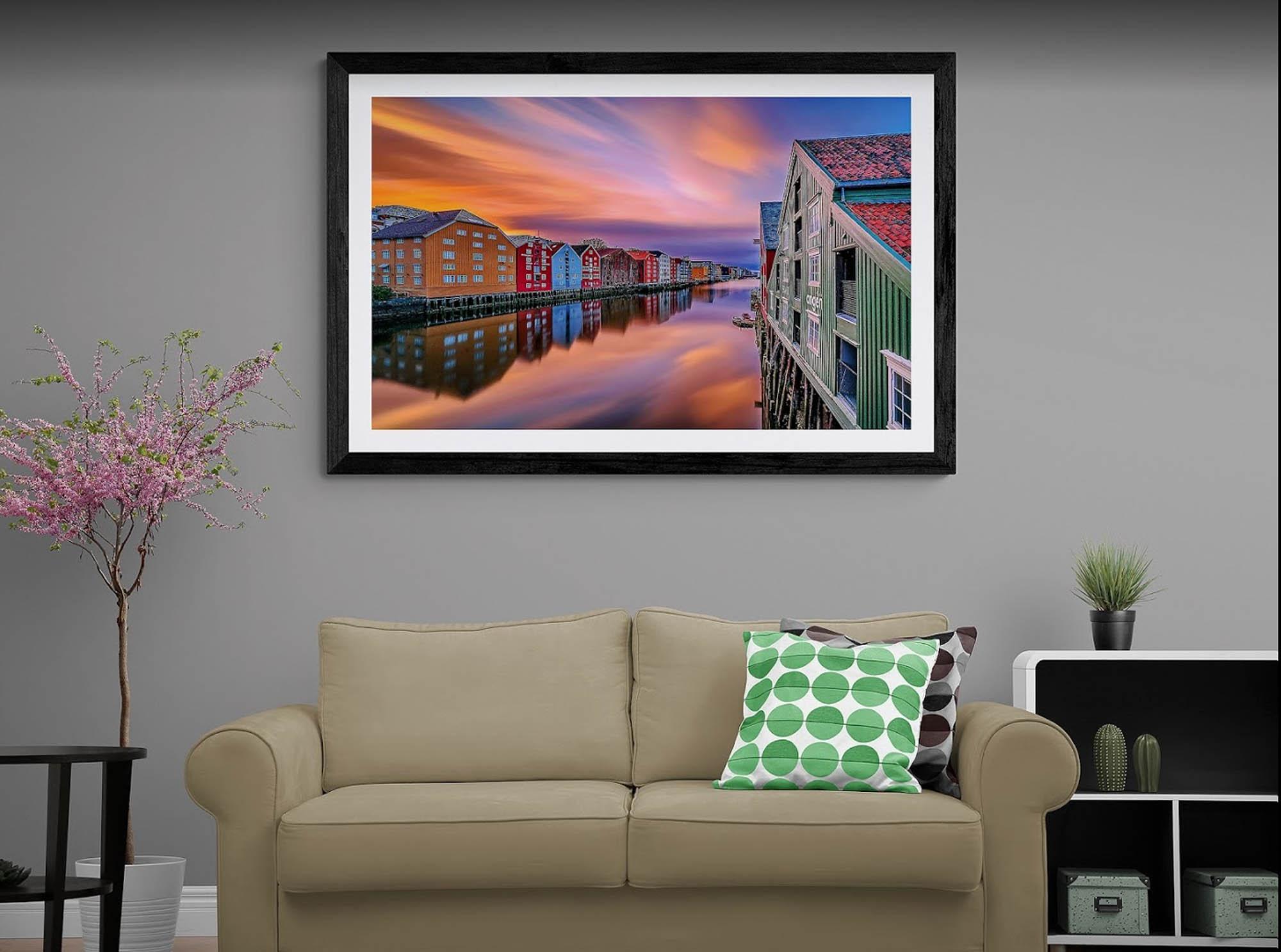 Et moderne stueinteriør med en sofa, et bilde av en solnedgang over en by med fargerike bygninger og vannrefleksjoner.