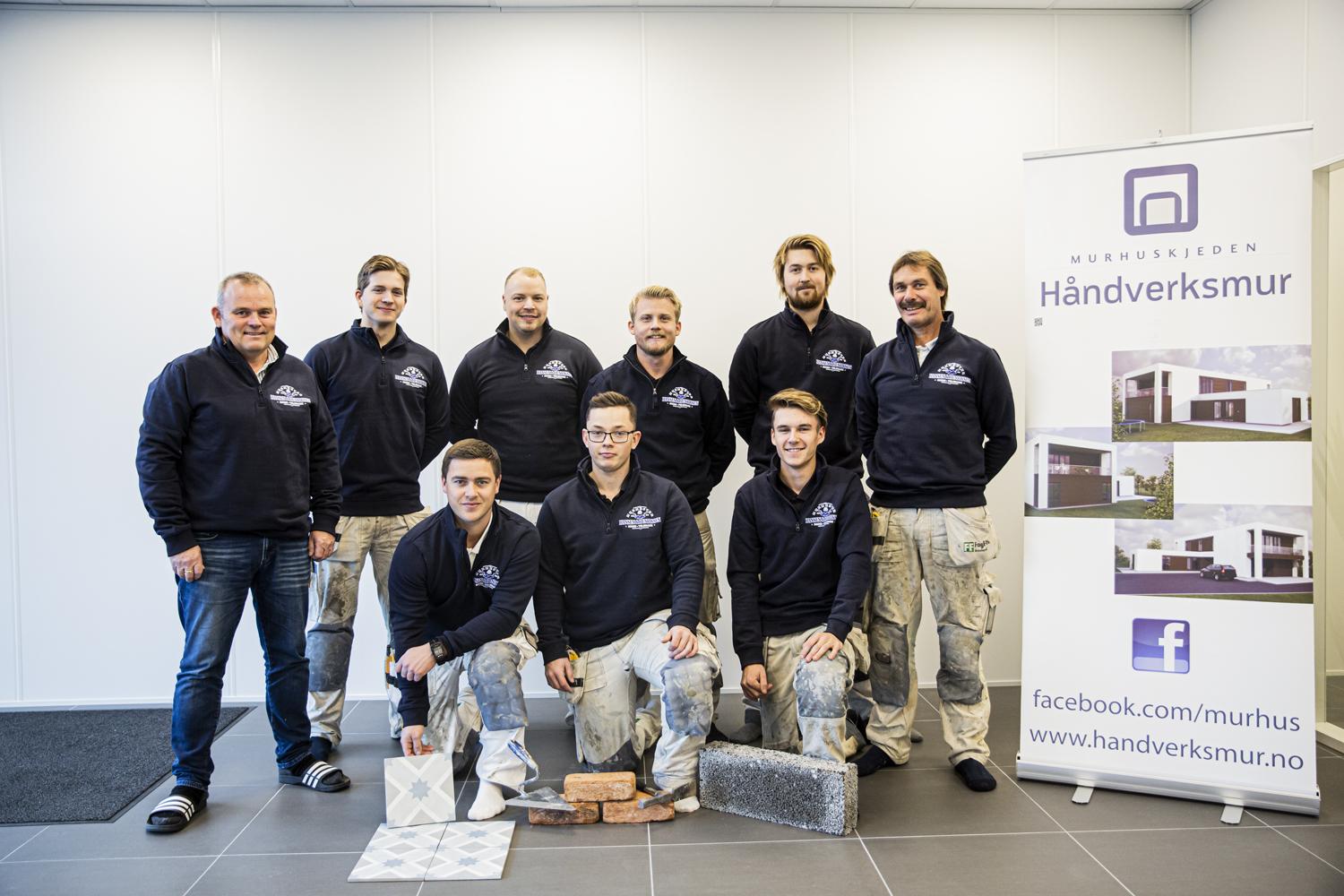Gruppebilde av håndverkere i arbeidsklær, poserer foran en banner for Håndverksmur.
