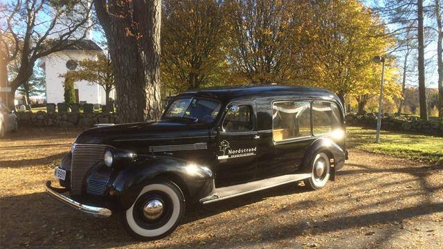 En svart veteranbil fra 1930-tallet parkert i en solfylt hage med trær i bakgrunnen.