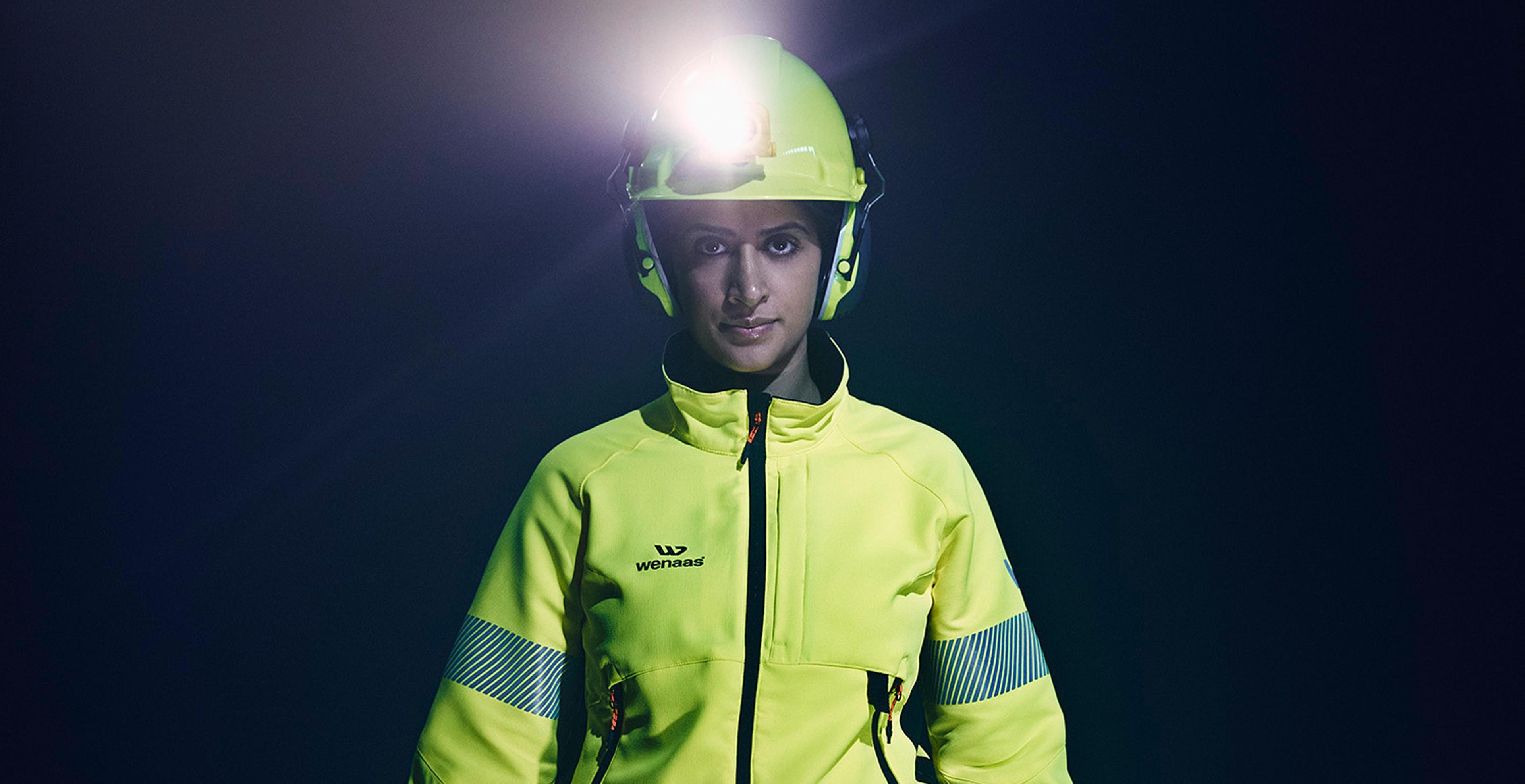 En person i gul sikkerhetsjakke og hjelm med hodelykt, står i mørket med et bestemt blikk.