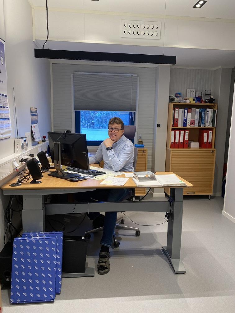 En mann sitter ved et skrivebord i et kontor, med datamaskin og dokumenter rundt seg.