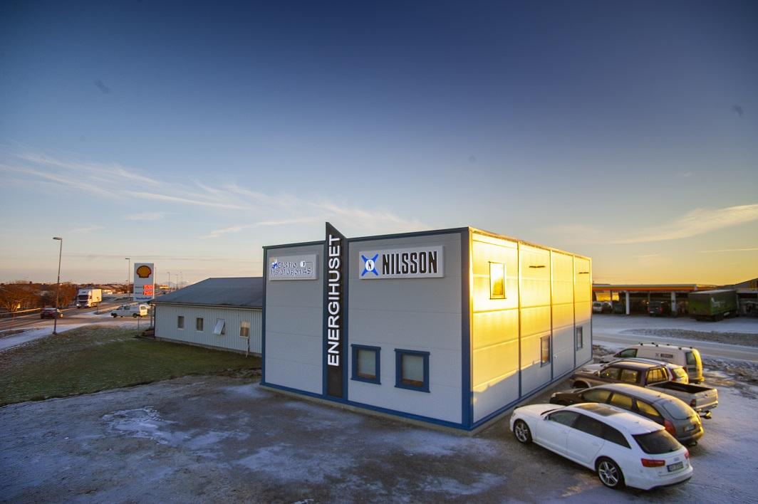 En moderne bygning med skilt for "Energihuset" og "Nilsson", omgitt av parkerte biler og en klar himmel.