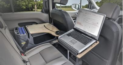 En laptop på et sammenleggbart bord i baksetet av en bil, med grå seter og en ryddig interiør.