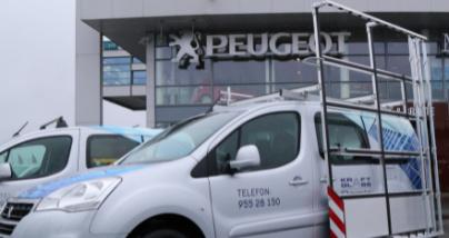 En grå Peugeot varebil parkert foran en Peugeot-forhandler med logo og telefonnummer synlig.