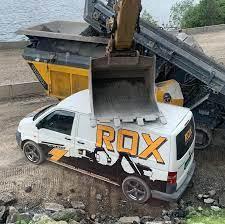 En hvit varebil med "ROX" på siden står under en gravemaskin som tømmer materiale fra en lastebil.