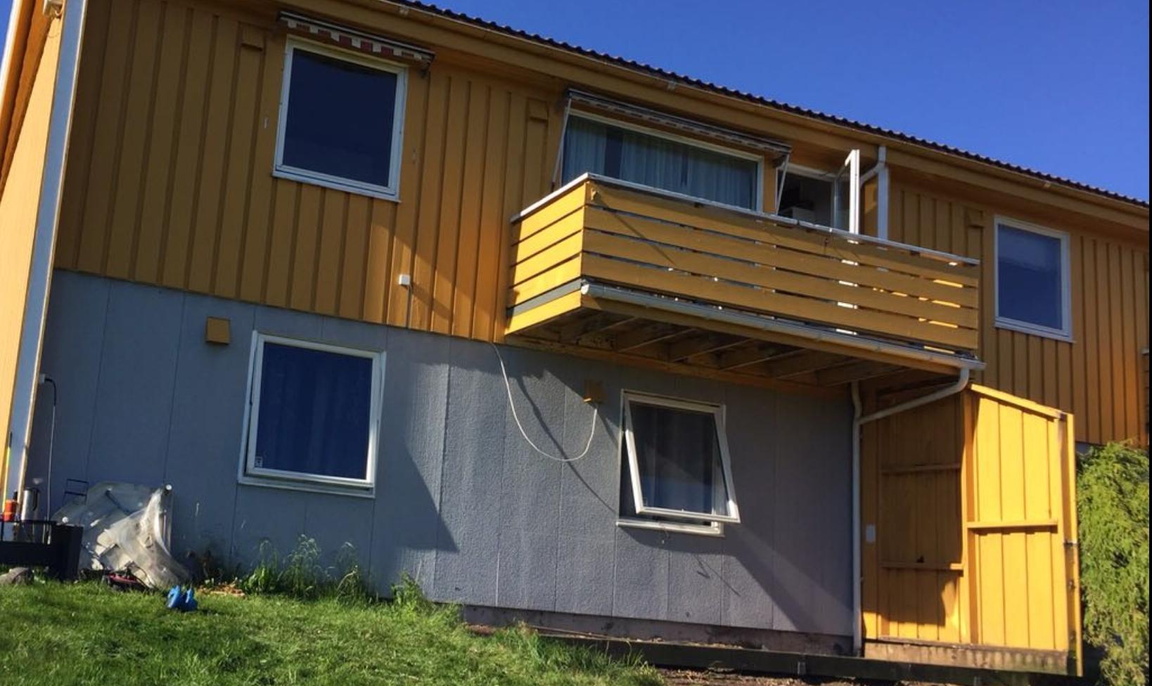 Gult hus med balkong og flere vinduer, plassert i en grønn hage under blå himmel.
