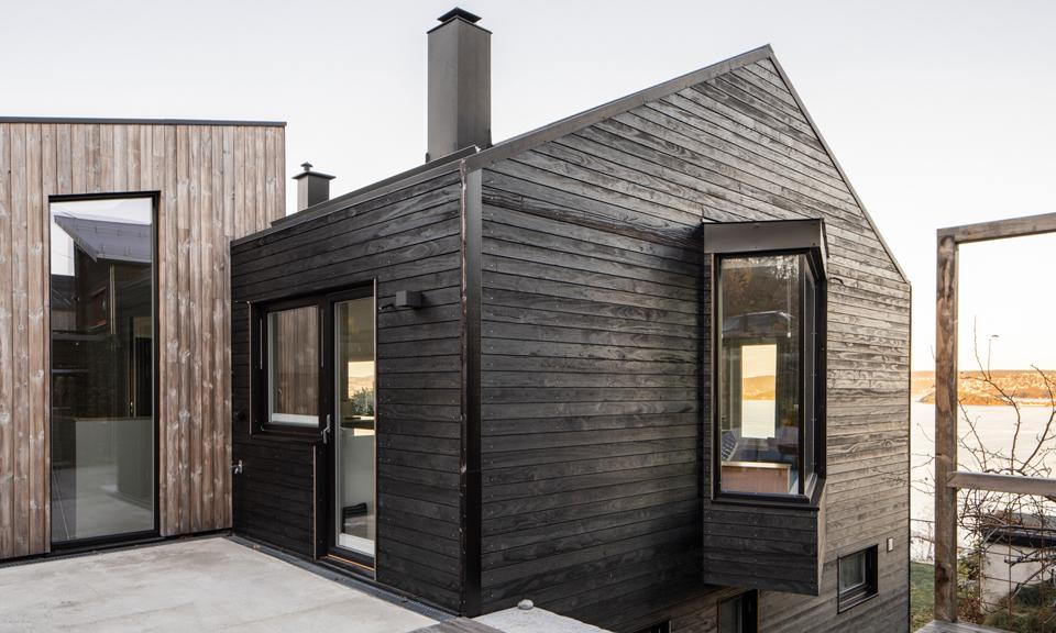 Moderne hus med svart trepanel og store vinduer, beliggende ved vannet.