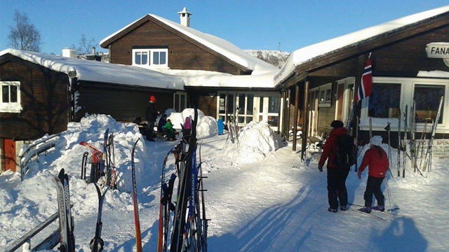 Folk går på snødekt vei foran en hytte med skiutstyr og snø i solfylt vintervær.