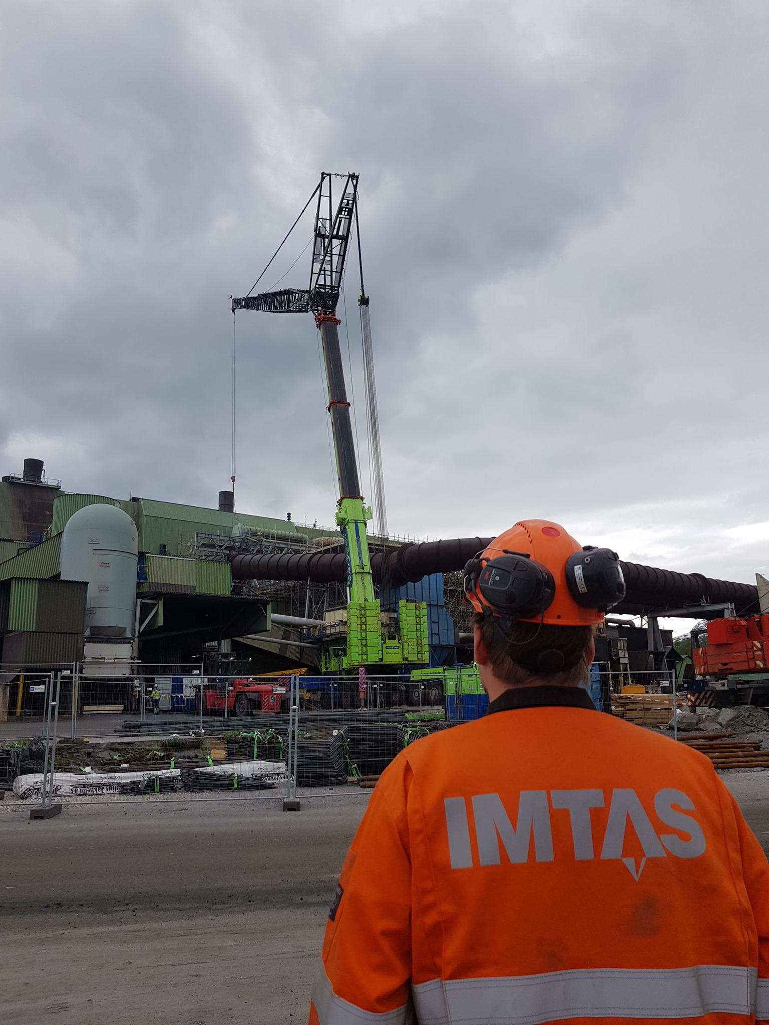 En arbeidstaker i oransje vernesjakke ser opp på en stor kran som løfter materialer på en byggeplass.