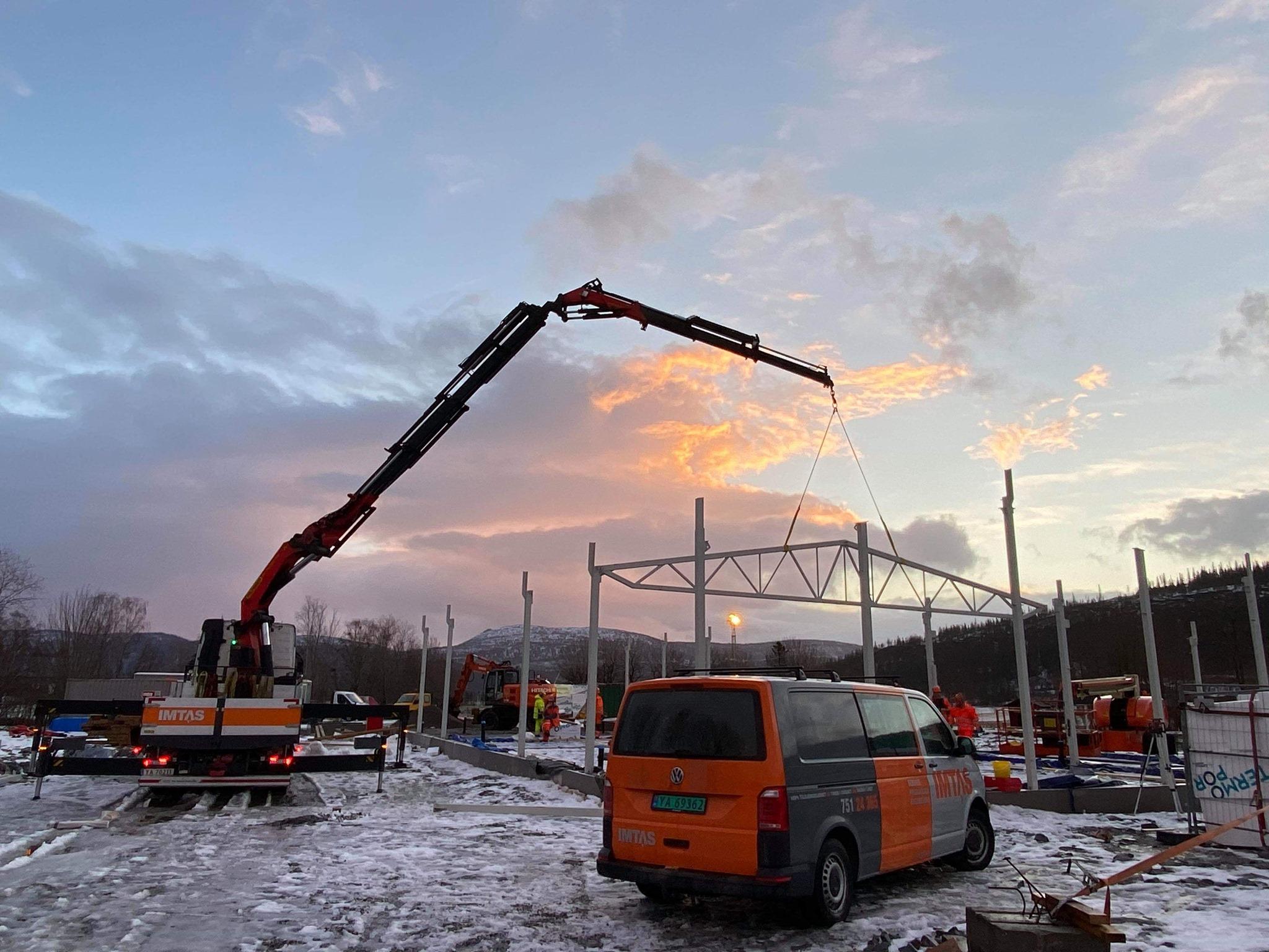 Kran løfter stålbjelker på byggeplass med solnedgang i bakgrunnen og en varebil parkert i forgrunnen.