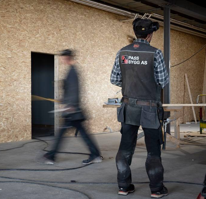En håndverker i arbeidsklær står foran en uferdig vegg, mens en kollega går forbi i bakgrunnen.
