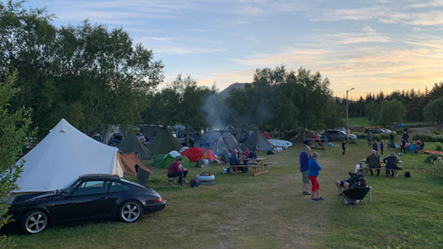 Campingplass med flere telt, folk som samles rundt bål, og biler parkert i naturskjønt landskap.