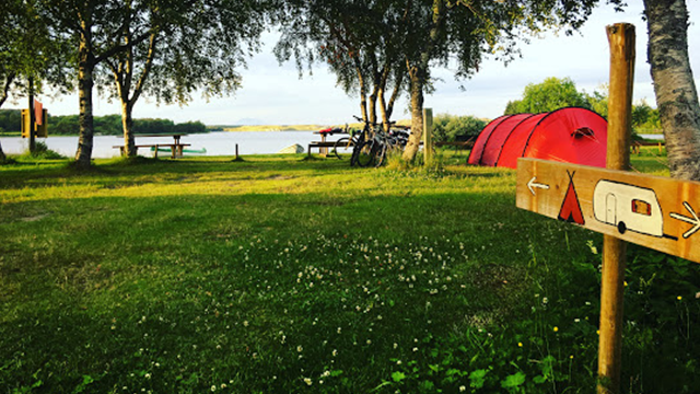 Campingplass ved vannet med telt, benker og en skilt som viser vei til campingvogn. Grønt gress og trær rundt.