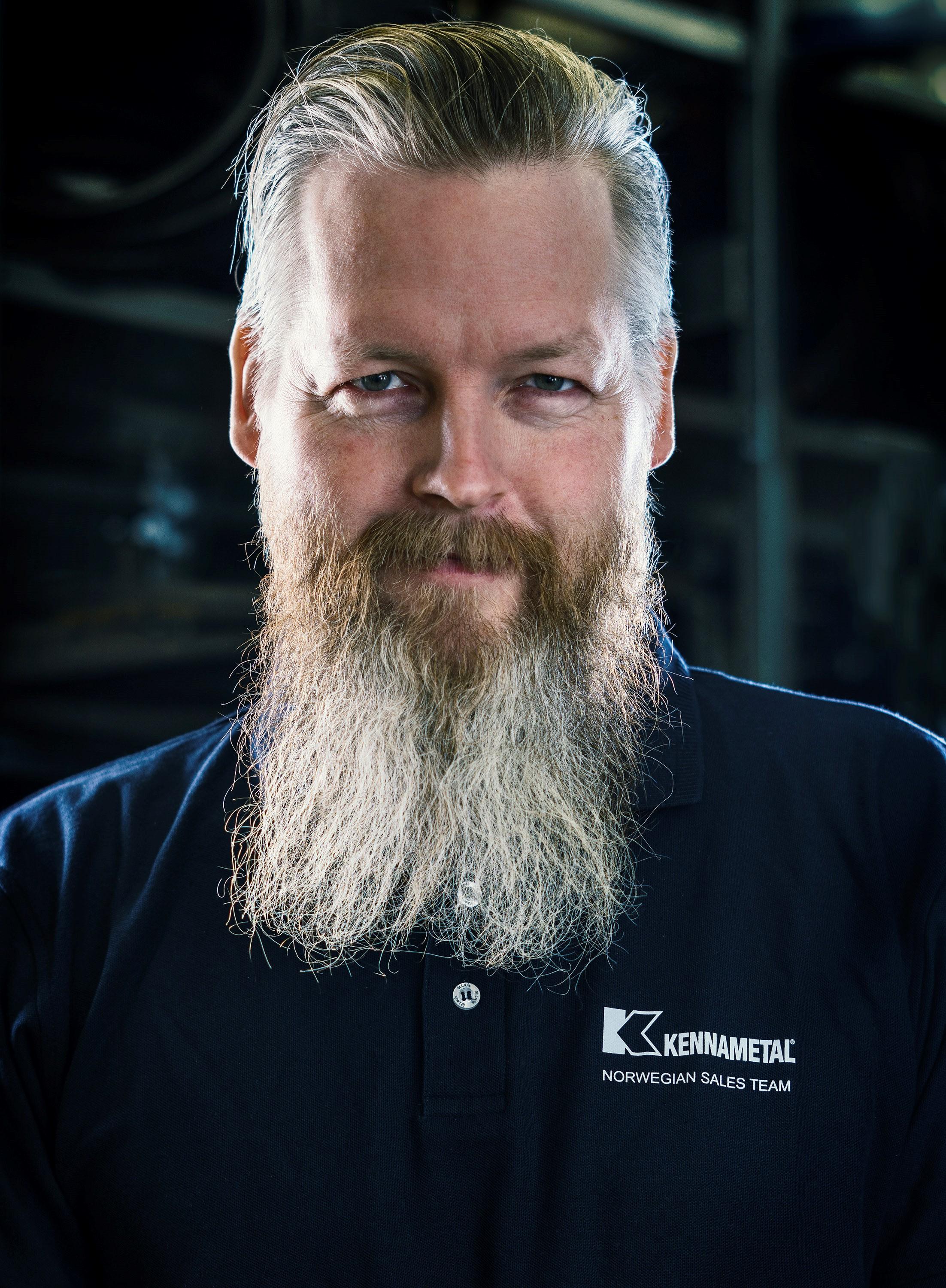 Mann med skjegg iført en mørk skjorte med logoen til Kebalmetal, smilende i en industriell setting.