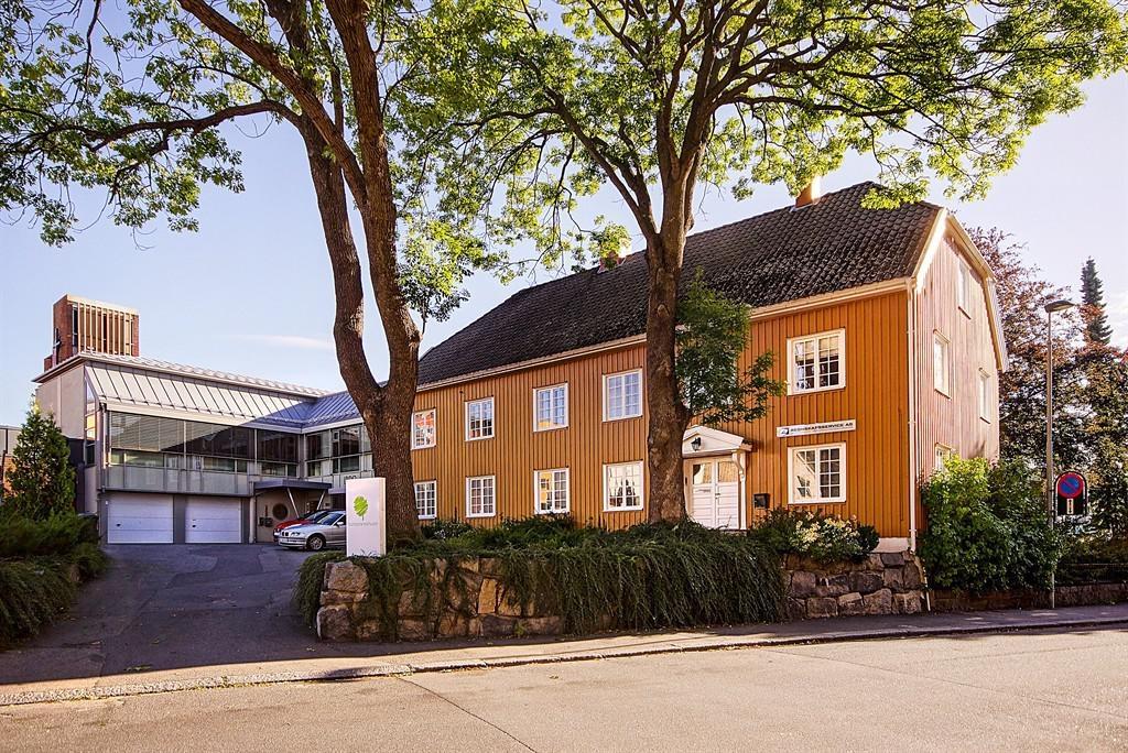 En stor, oransje trebygning med hvite vinduer, omgitt av trær og en moderne bygning i bakgrunnen.