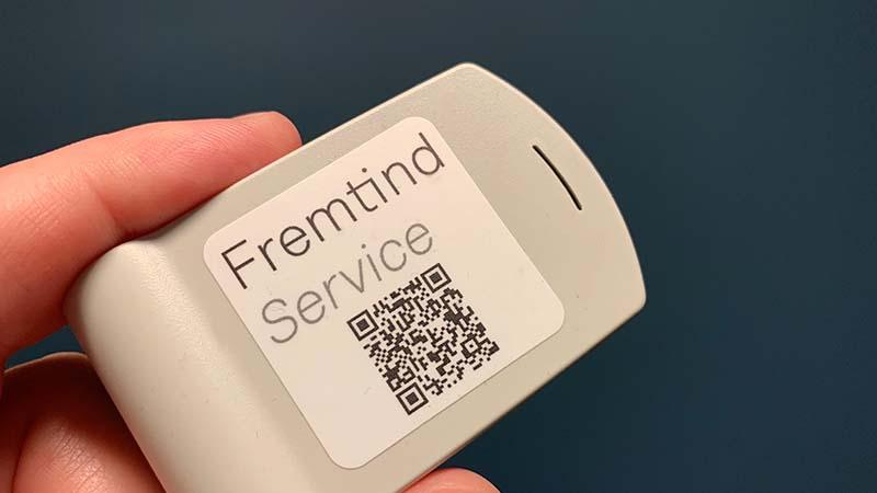 En hånd som holder en grå enhet med Fremtind Service og en QR-kode på etiketten.