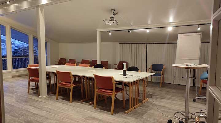 Møterom med lang bord, stoler og prosjektor, beliggende i et lyst rom med store vinduer.