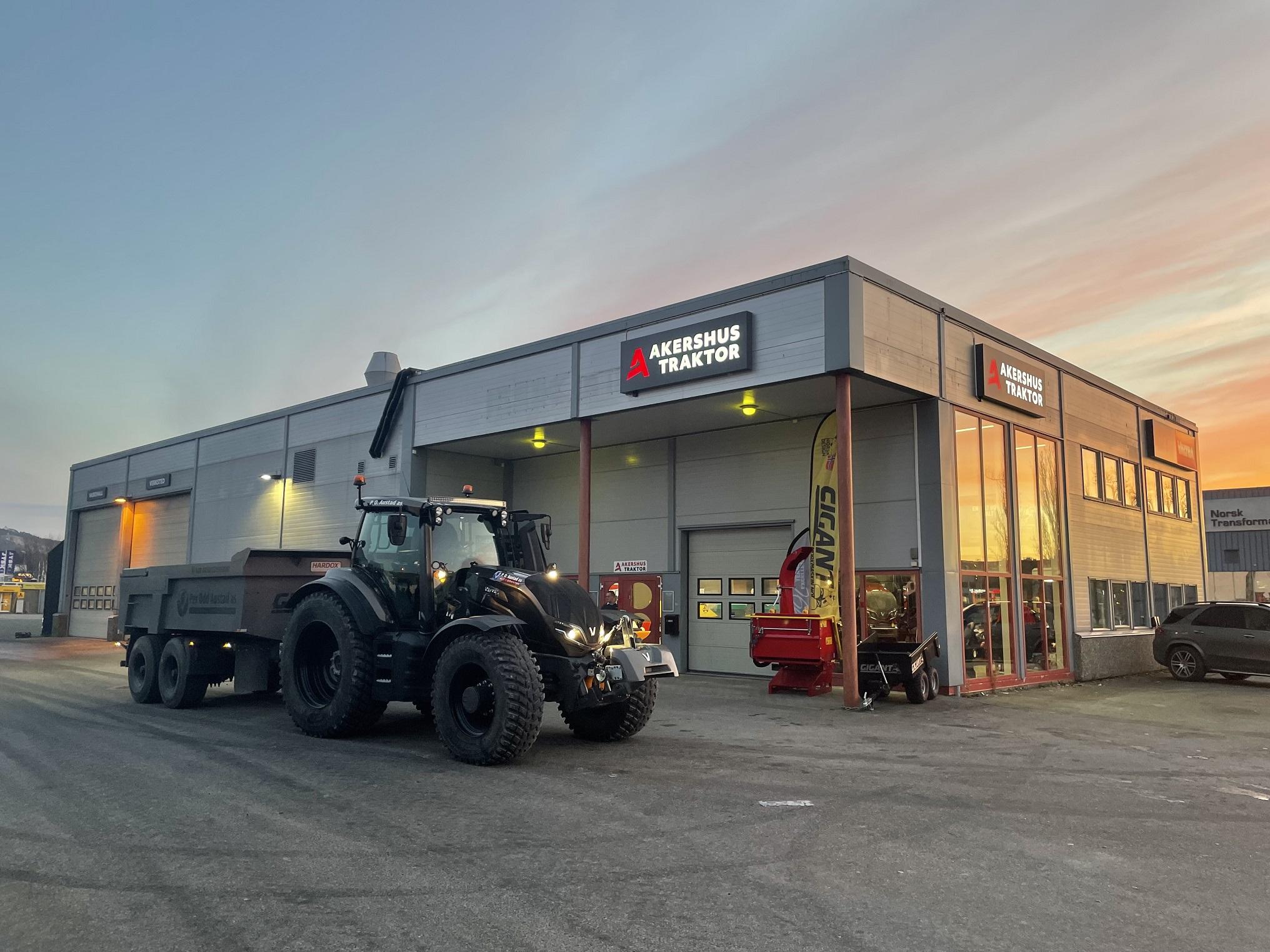 En stor traktor med tilhenger står foran en moderne bygning med skiltet "Bergsjø Traktor" i solnedgang.