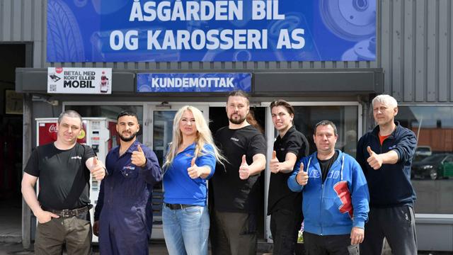 En gruppe ansatte ved Åsgarden Bil og Karosseri AS står foran bygningen og gir tommel opp.