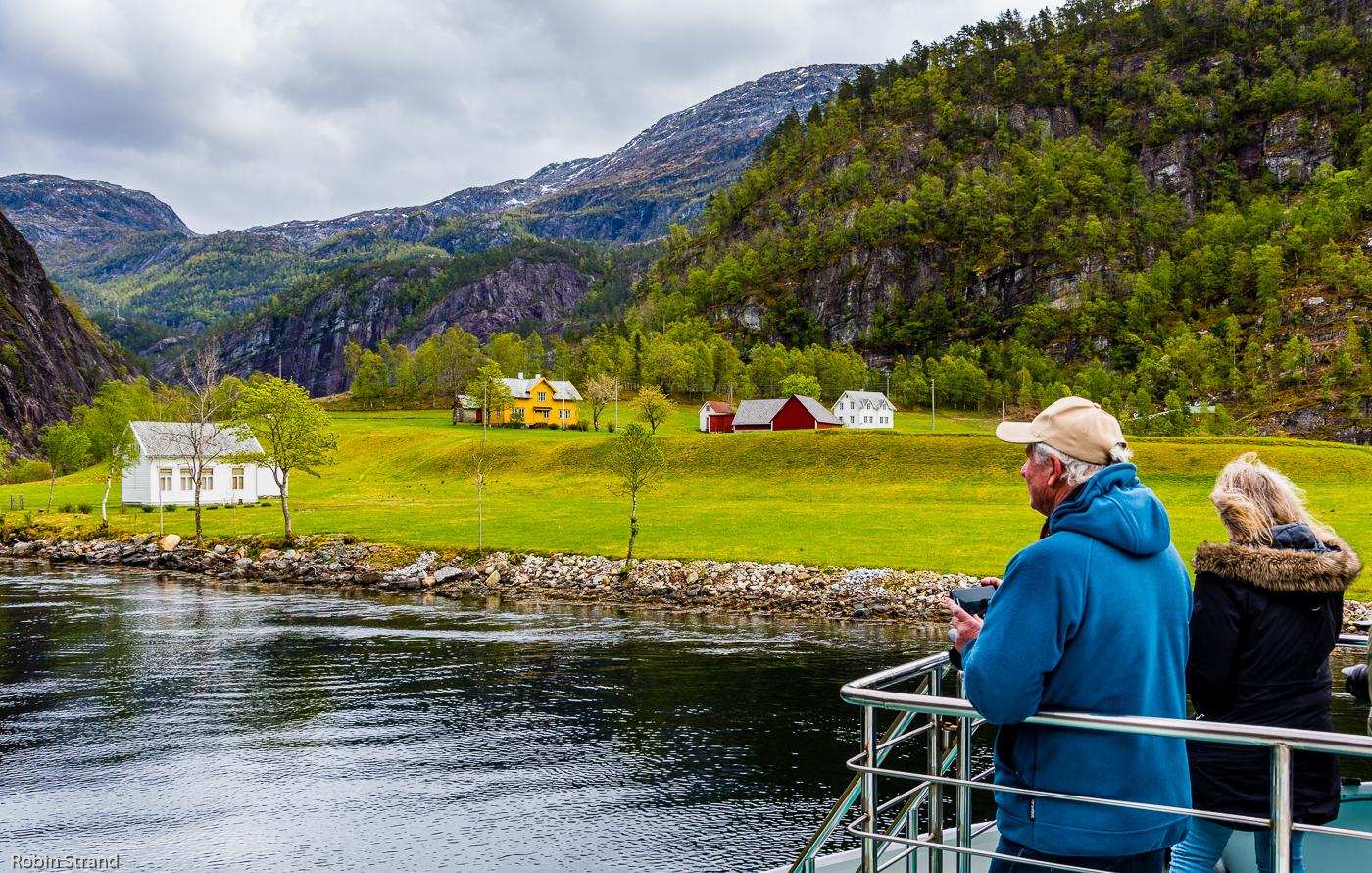 To personer står på en båt og ser på landskapet med grønne åser og fargerike hus ved fjorden.