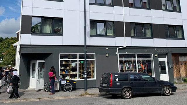 En gateutsikt av en moderne bygning med butikkvinduer, en svart bil parkert og folk som går forbi.