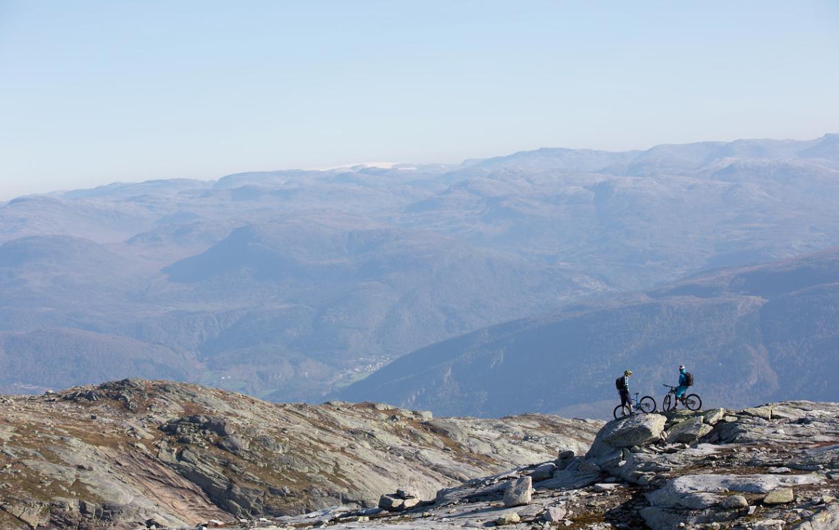 To personer på terrengsykler som står på en steinete fjelltopp med fjellandskap i bakgrunnen.