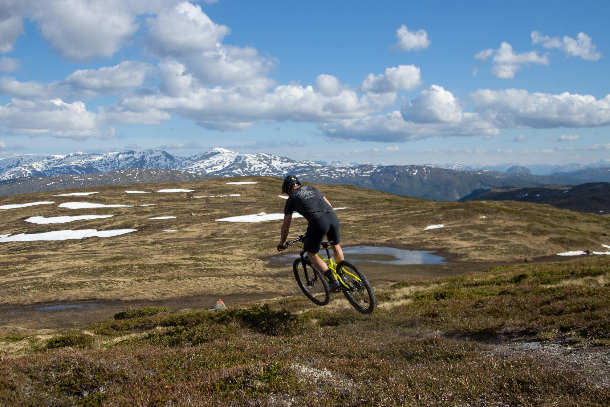 En syklist som hopper over en bakke i fjellandskap med snø og fjell i bakgrunnen.