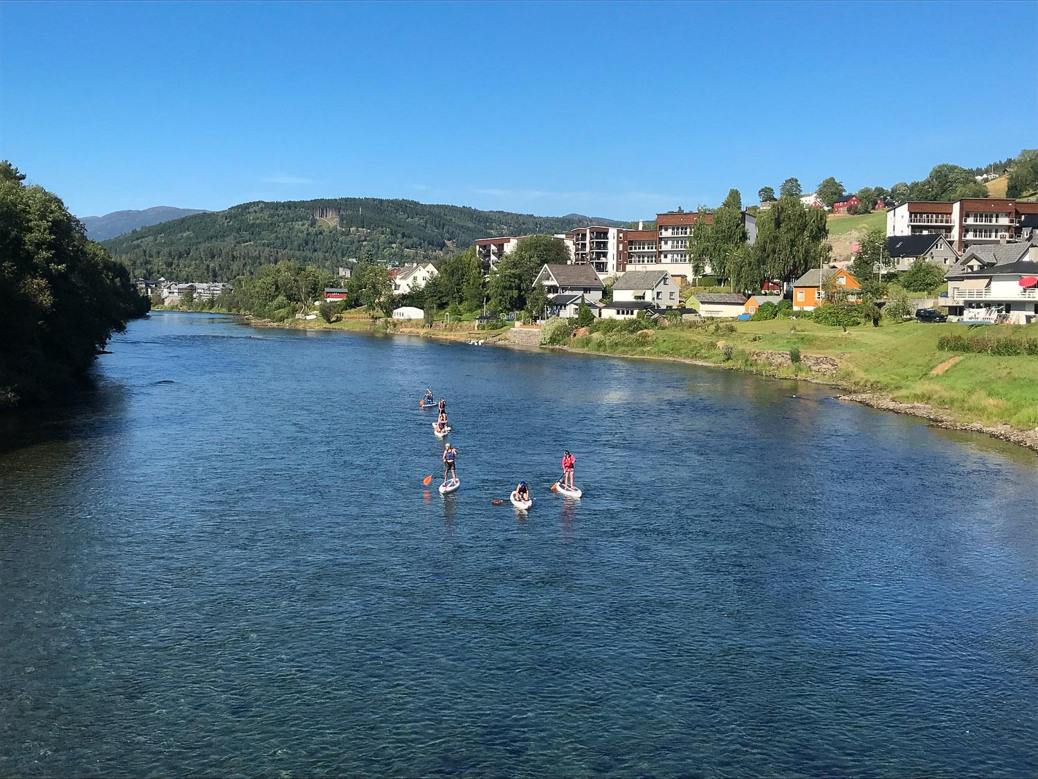 Folk som padler på elven med hus og fjell i bakgrunnen en solfylt dag.