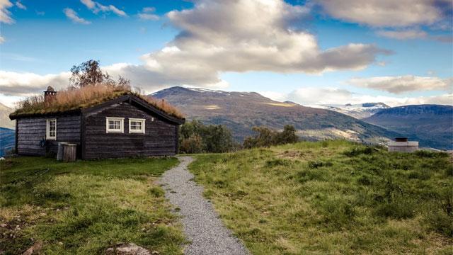 En koselig hytte med gressdekt tak, omgitt av fjell og grønt landskap under en blå himmel.