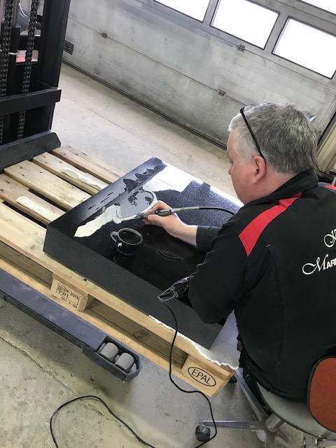 En mann sitter på en stol og arbeider med en svart gjenstand på en pall i et verksted.