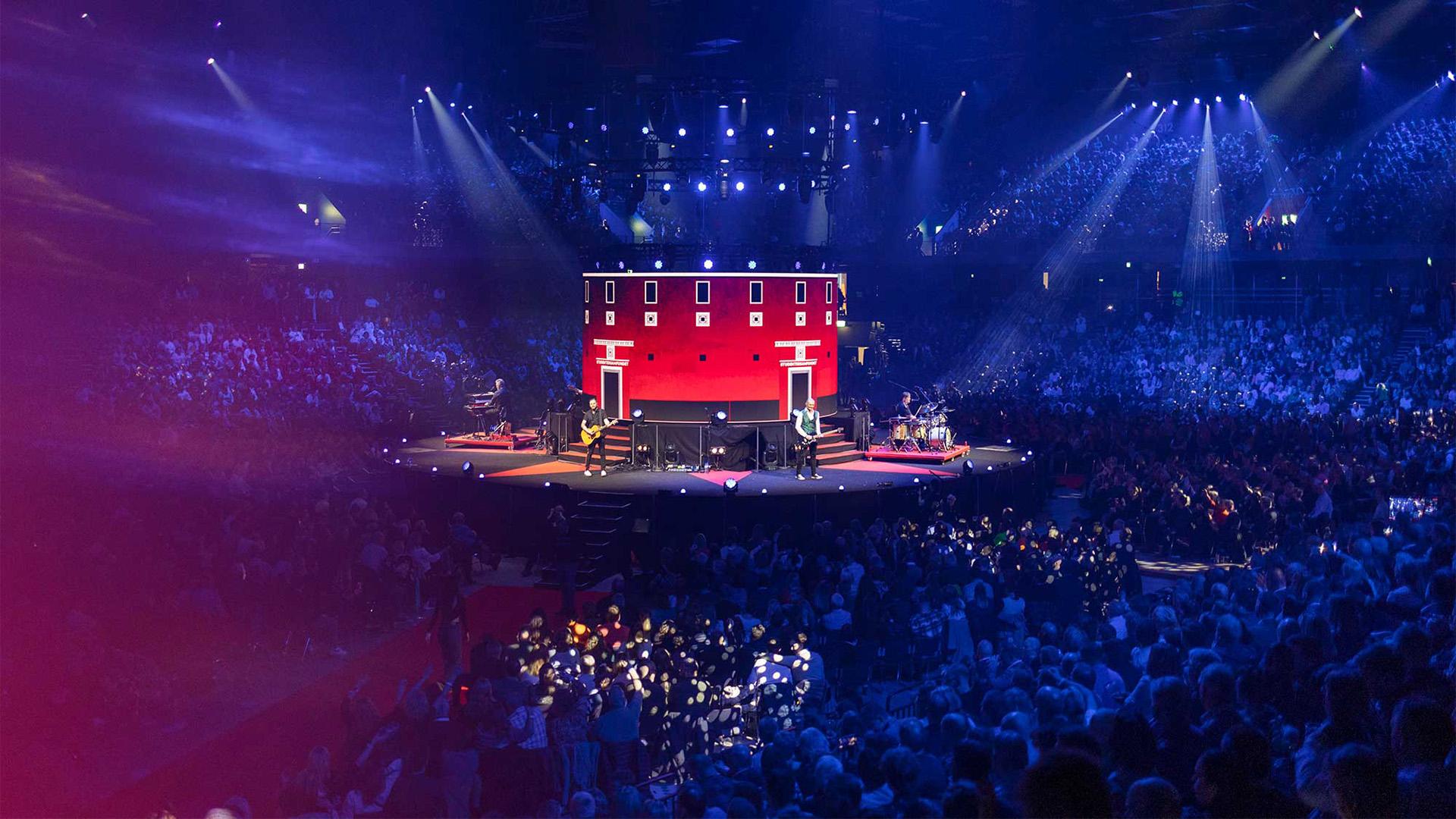 En stor konsert med lys og en stor scene, omgitt av et entusiastisk publikum i en fylt arena.