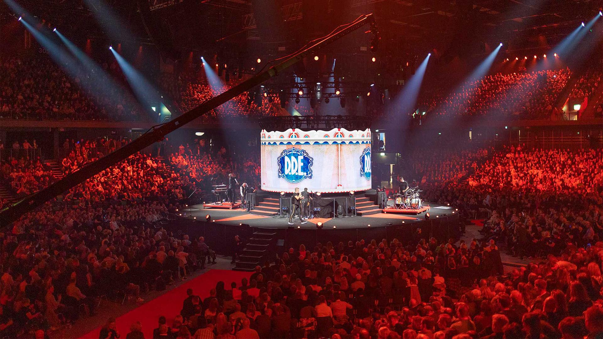 En stor konsert med mange mennesker i publikum, lys og en scene med musikere som spiller.