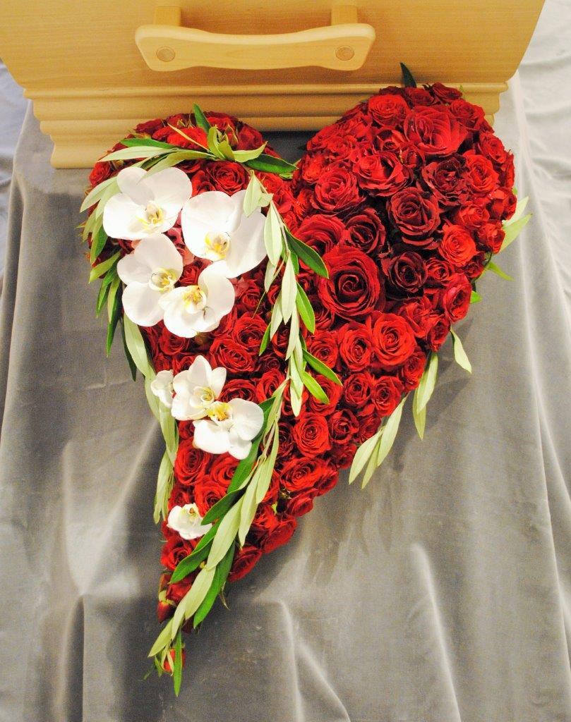 Et hjerteformet blomsterarrangement laget av røde roser og hvite orkideer, plassert på en grå duk.