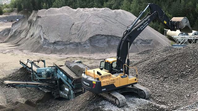 Gravemaskin og knusemaskin jobber på en grusplass med store haug av grus og sand i bakgrunnen.