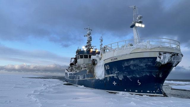 Fiskebåt som ligger fast i isen, med snø og frost på dekk under en grå himmel.