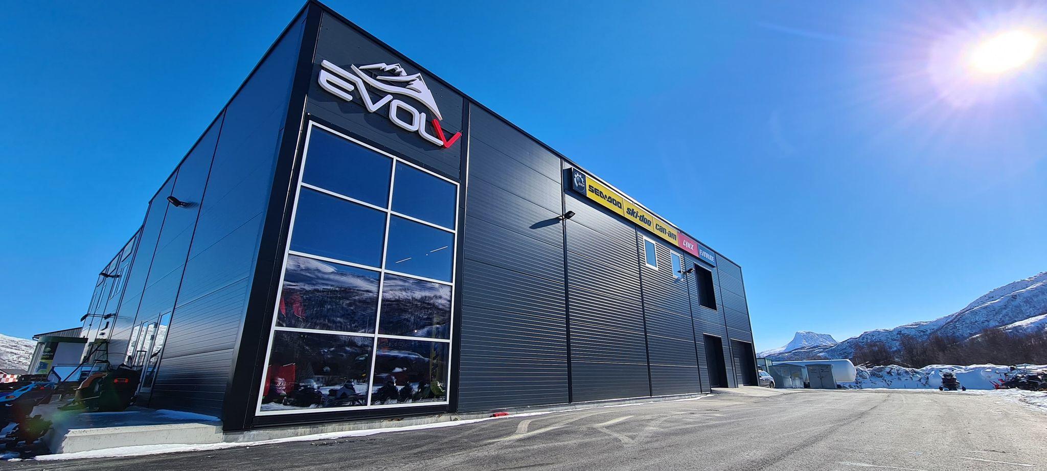 Moderne bygning med store vinduer, merker som EVO og andre, omgitt av snødekte fjell under klar blå himmel.