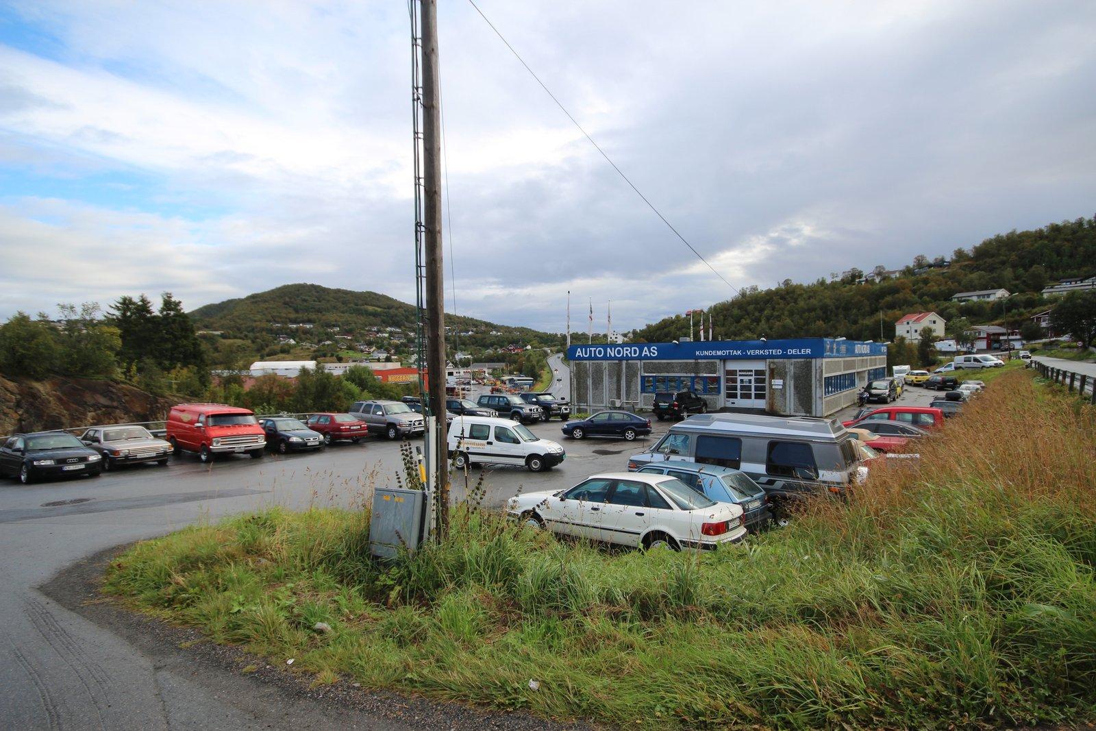 Bilde av en bilforhandler med flere biler parkert og en bakgrunn av grønne åser under en skyet himmel.