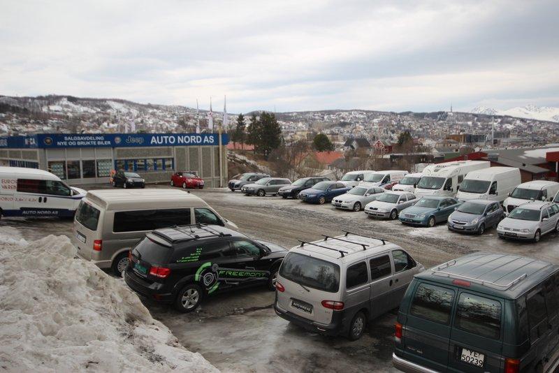 Bilde av en bilforhandler med flere biler parkert på snødekt område, med fjell i bakgrunnen.