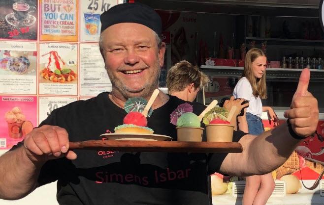 En smilende mann med en hatt holder en brett med iskremer foran en iskiosk.