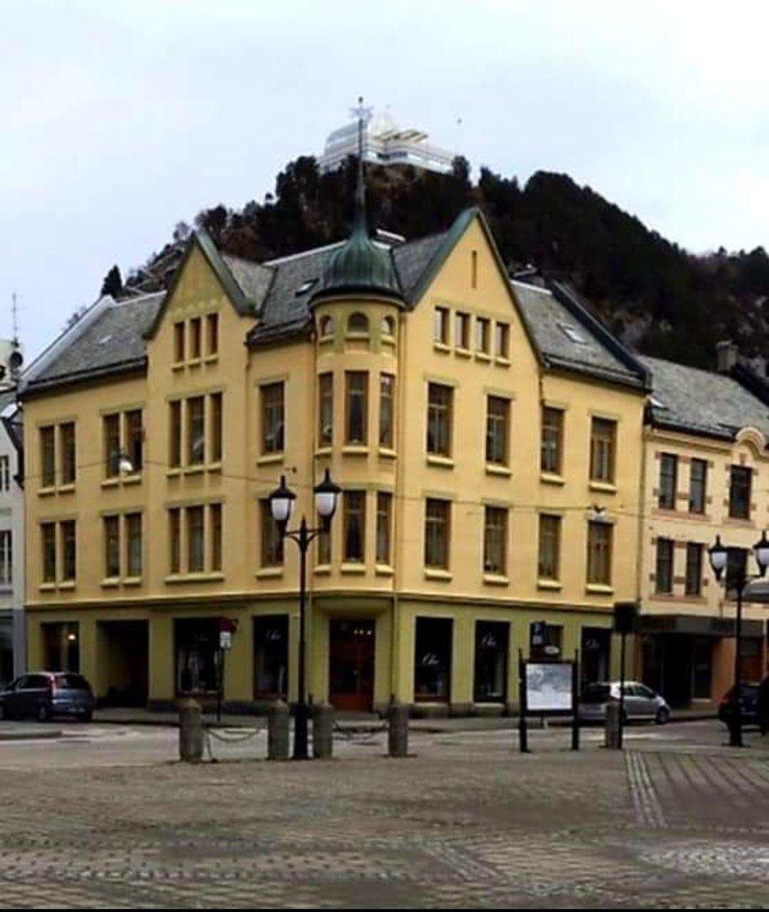 Gul bygning med tårn og grønne detaljer på en plass, med fjell i bakgrunnen og biler parkert.