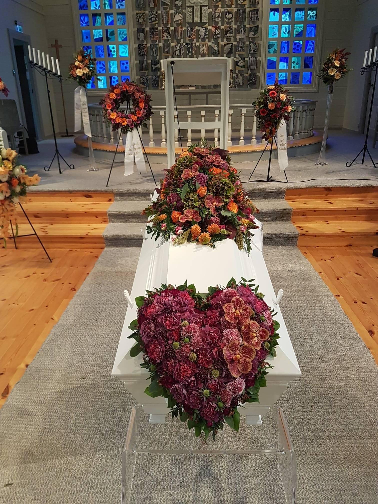 Kiste dekorert med blomster i hjerteform, omgitt av blomsteroppsatser i en kirke.