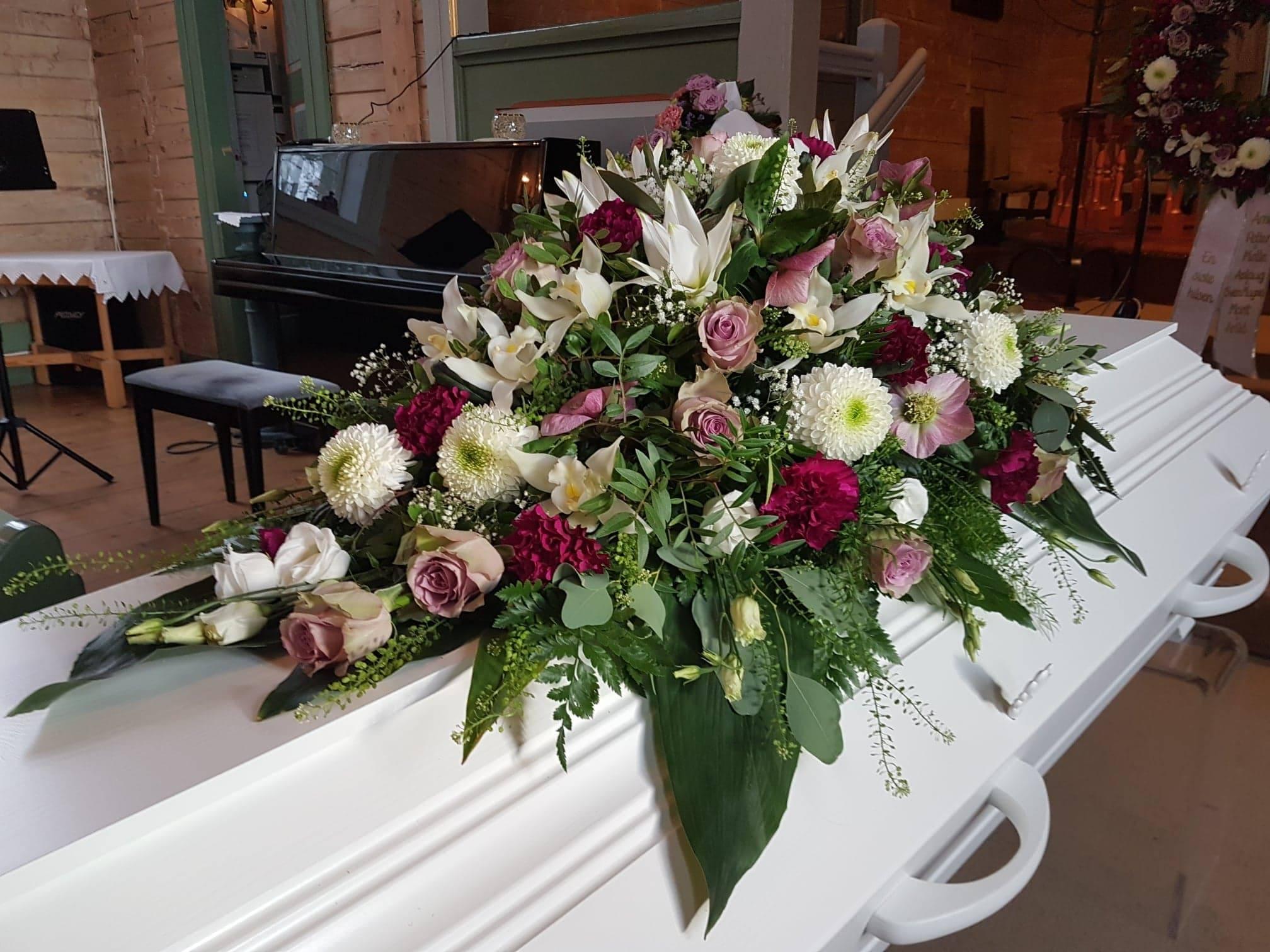 En hvit kiste dekorert med en vakker blomsterkrans av liljer, roser og andre blomster i ulike farger.