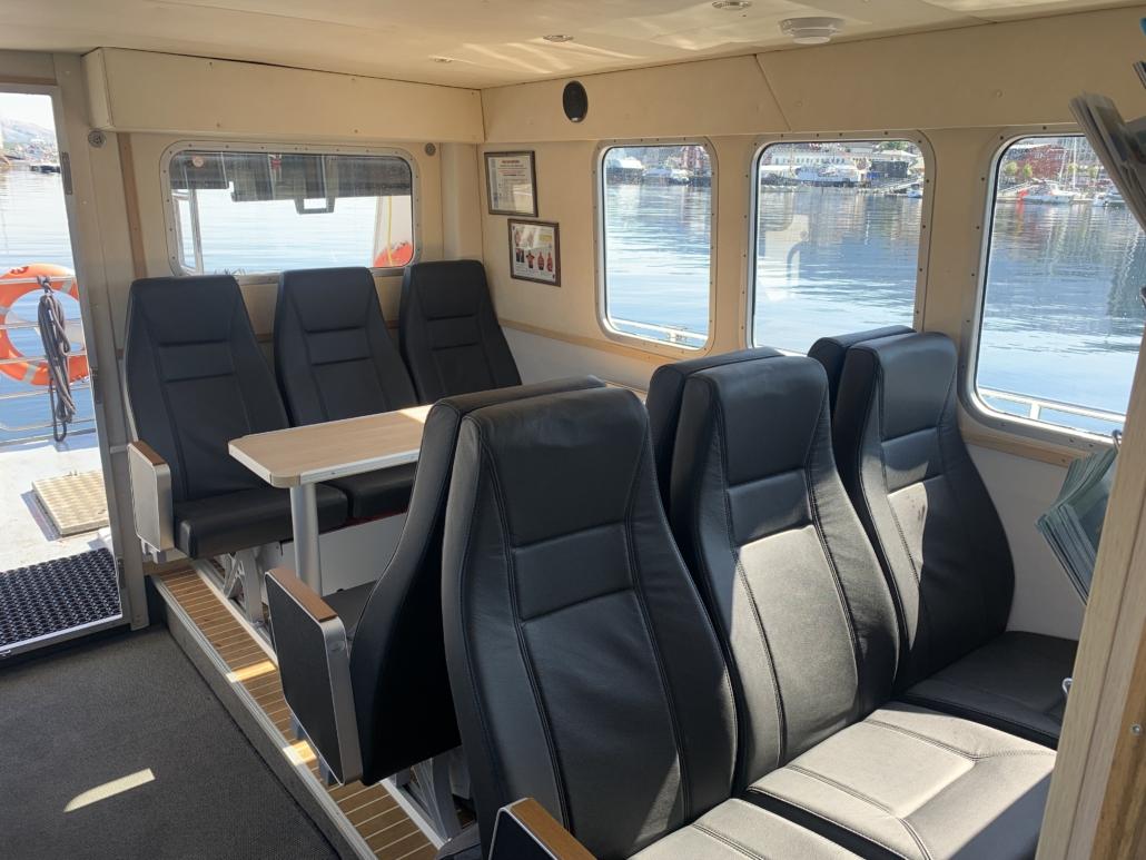 Innvendig bilde av en båt med komfortable seter og bord, med utsikt mot vannet og havnen.