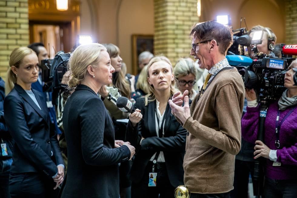 En kvinne i svart jakke snakker med en mann i brunt, omgitt av journalister og kameraer i en offentlig setting.