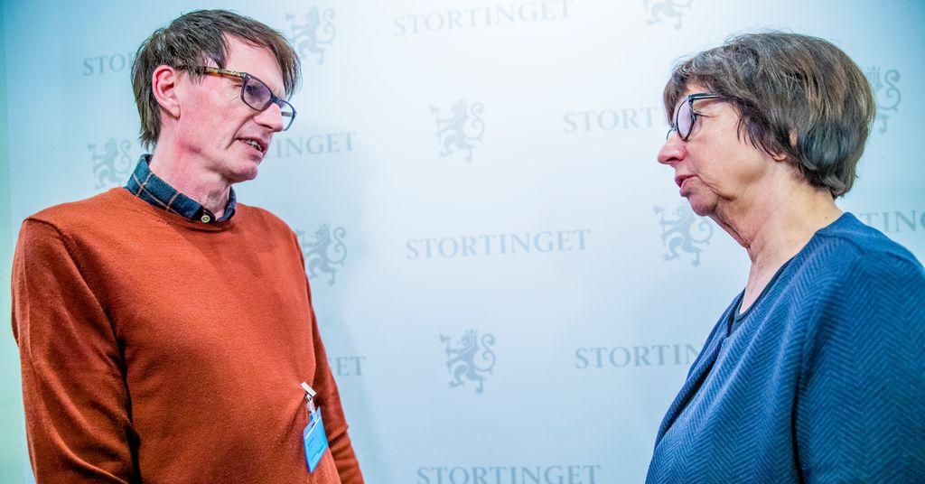 To personer i samtale foran en bakgrunn med Stortingets logo. Den ene har på seg en oransje genser.